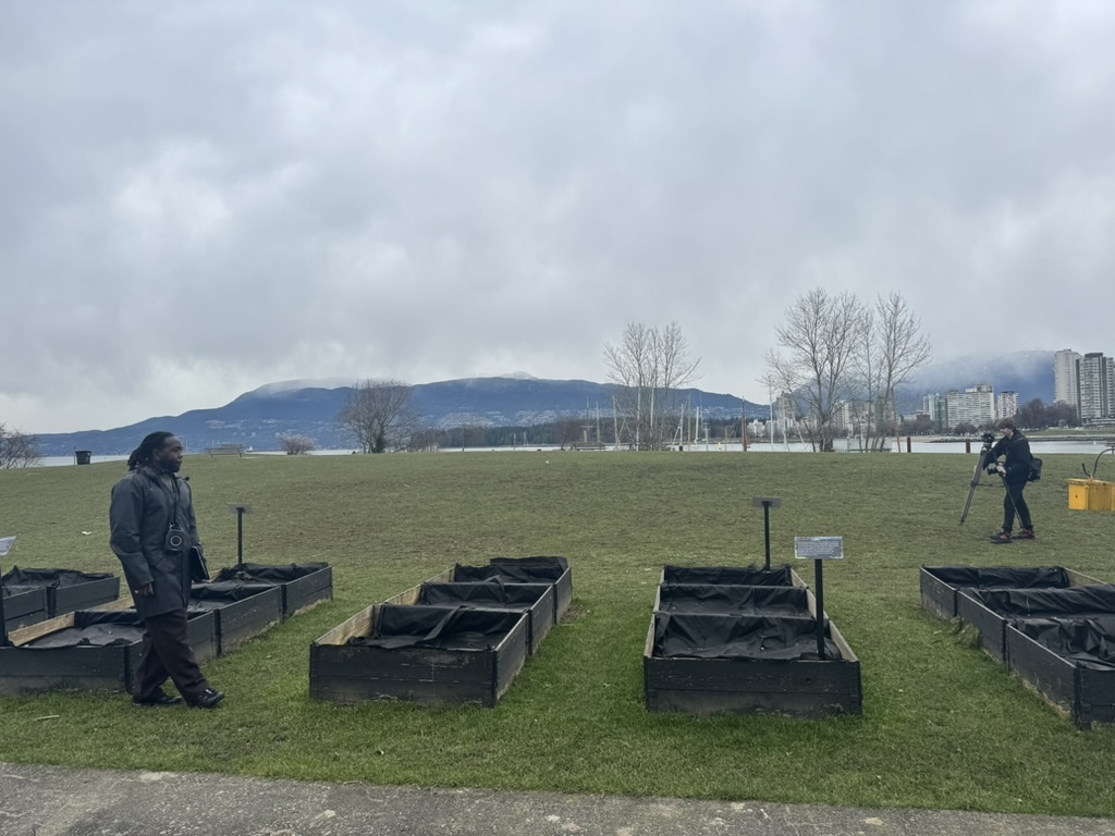 Kor the talk host standing in front of some empty garden boxes out front a grassy field on the ocean shore