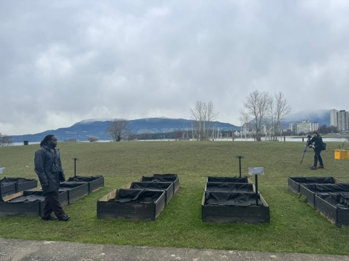 Kor the talk host standing in front of some empty garden boxes out front a grassy field on the ocean shore